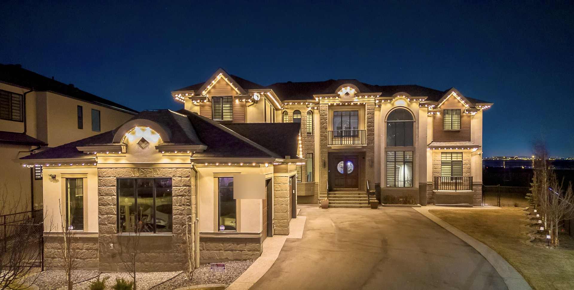 The same stone-and-stucco home seen from the driveway at blue hour, warm white light following the roofline across both wings.