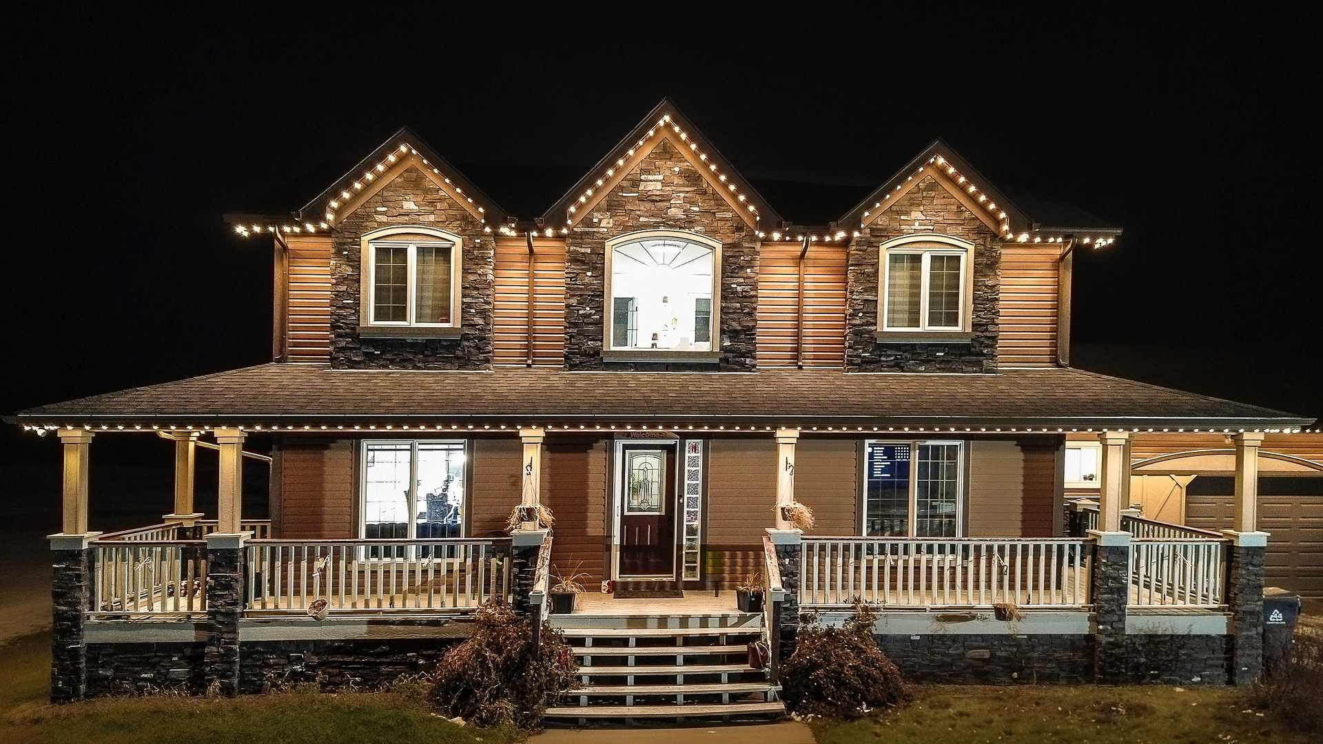 An Alberta two-storey home at night, its gables, eaves and wraparound porch traced in even warm-white light.