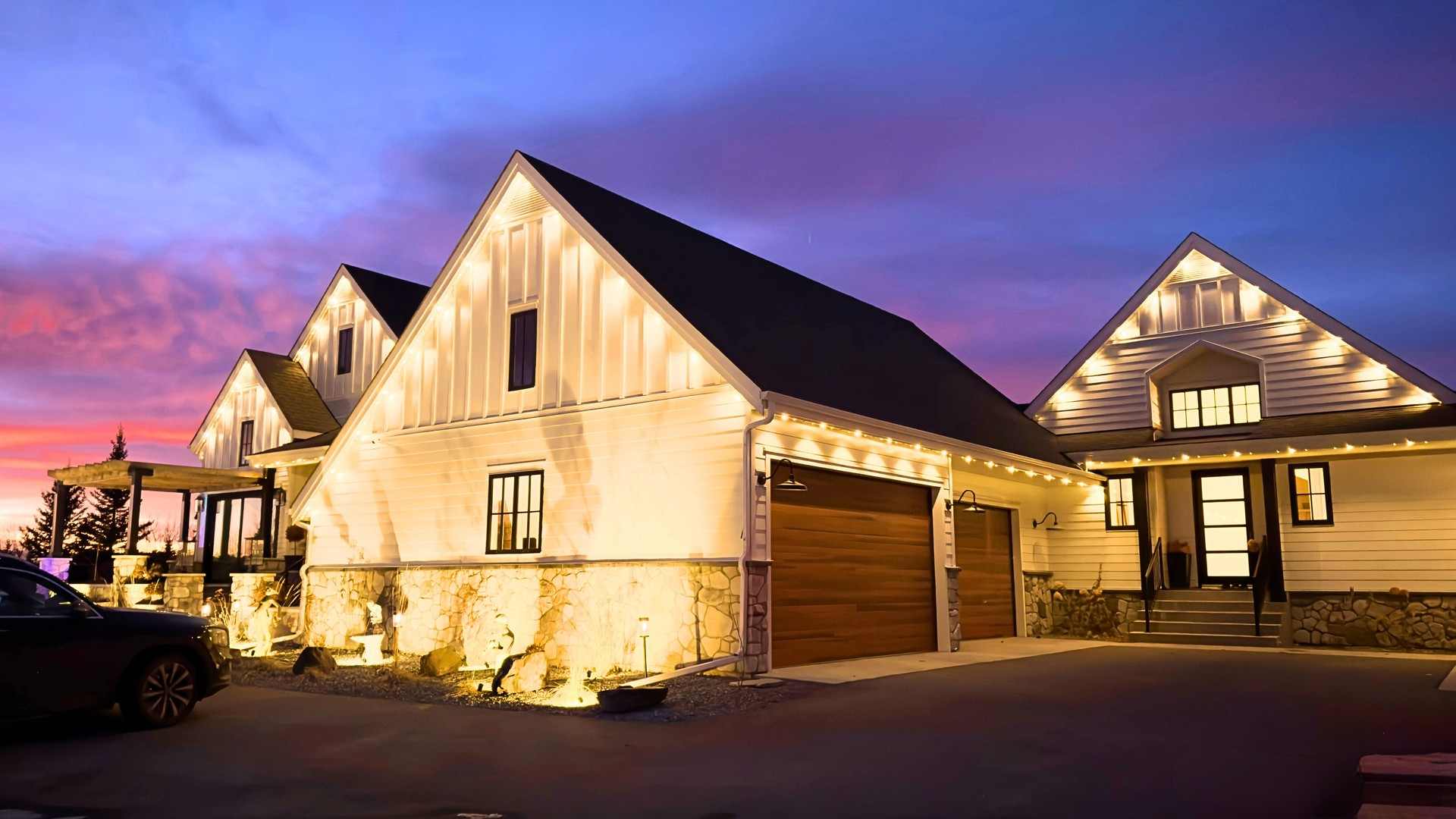 A white modern farmhouse at sunset, its steep gables and garage lit in warm white against a pink and violet Alberta sky.